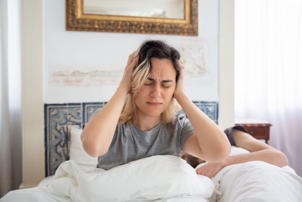 Mujer sentada en la cama por la mañana con las manos en la cabeza por dolor de cabeza al despertar