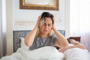 Mujer sentada en la cama por la mañana con las manos en la cabeza por dolor de cabeza al despertar
